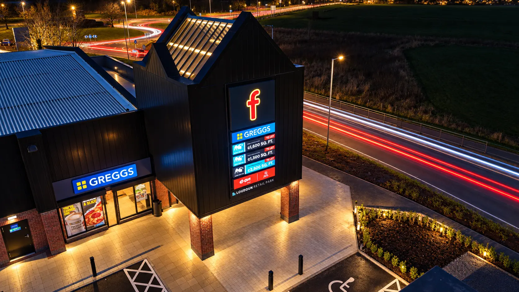 Aerial night view of Loudoun Retail Park beside the A71, with the illuminated Greggs store, tenant totem sign and traffic light trails