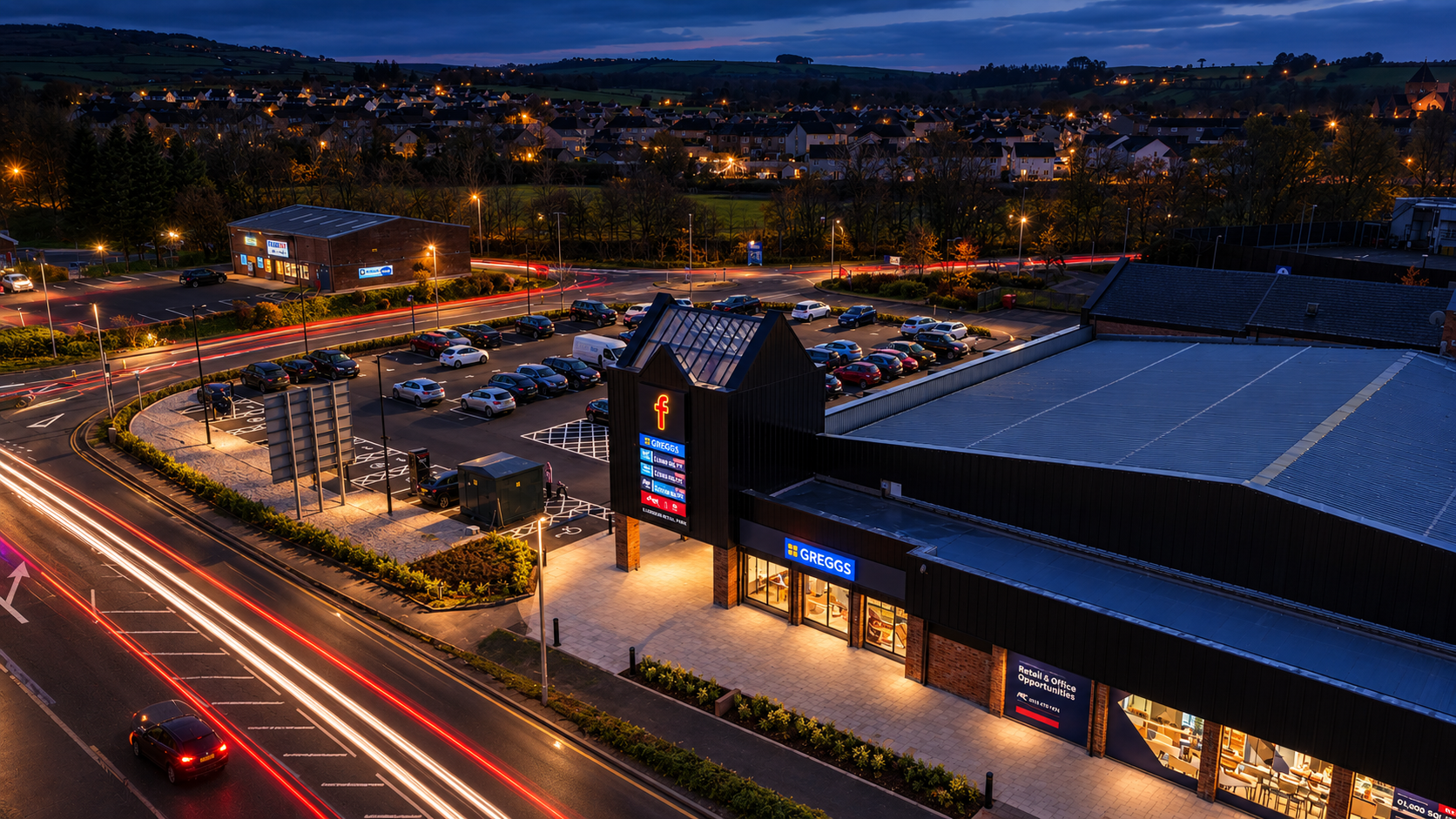 Loudoun Retail Park at dusk with light trails along the A71, showing the Greggs storefront, car park and tenant totem